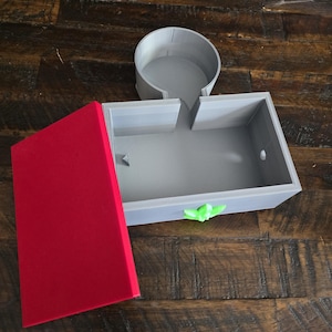 Könnte beinhalten: Eine graue, 3D-gedruckte Box mit rotem Deckel und einem grünen schmetterlingsförmigen Griff. Ein separater grauer, kreisförmiger Behälter ist ebenfalls vorhanden. Die Artikel werden auf einer dunklen Holzoberfläche präsentiert. Die Box scheint ein Aufbewahrungsbehälter zu sein.