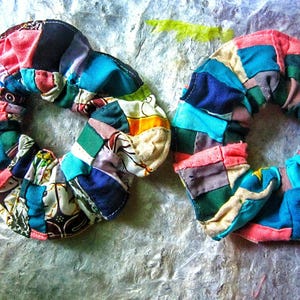 Pode incluir: Duas scrunchies coloridas em patchwork. Cada scrunchie é um anel de quadrados de tecido em várias cores, incluindo azul, rosa, verde e amarelo. As scrunchies são feitas à mão e têm uma estética vintage ou boêmia.
