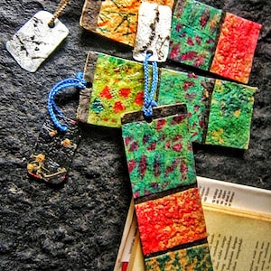 May include: Handmade bookmarks with vibrant, textured designs in green, red, orange, and brown. The bookmarks have a variety of patterns and are attached to small tags with cords.
