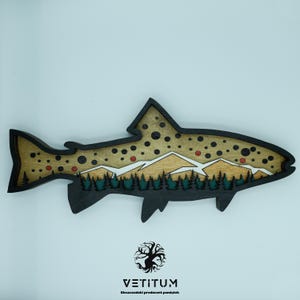 May include: Wooden fish-shaped wall art with a mountain and forest scene. The fish is brown with black outlines and features a detailed landscape with trees, mountains, and red and black dots. The art piece is a decorative item.