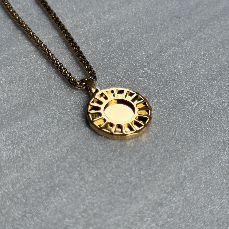 May include: Gold-toned necklace featuring a circular pendant with a sunburst design. The pendant has a central, smooth surface surrounded by a geometric pattern. The chain is a wheat-style chain. The necklace is displayed on a gray surface.