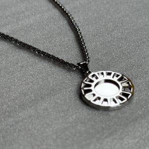 May include: A silver-toned necklace with a circular pendant. The pendant has an oval-shaped center and a decorative, openwork design around the edge. The chain is a fine, textured style, and the background is a solid gray.