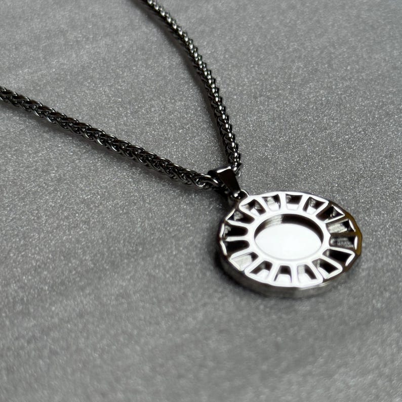 May include: A silver-toned necklace with a circular pendant. The pendant has an oval-shaped center and a decorative, openwork design around the edge. The chain is a fine, textured style, and the background is a solid gray.