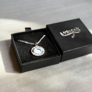 Op de afbeelding: Een zilverkleurige hangerketting met een cirkelvormig ontwerp, gepresenteerd in een open zwarte doos. Op het deksel van de doos staat de merknaam "LYRICAL". De ketting heeft een ketting en een gedetailleerde hanger. De doos is gevoerd met zwarte stof.