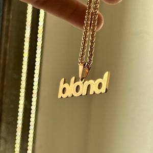 Op de afbeelding: Goudkleurige ketting met het woord "blond" in een gestileerd lettertype. De hanger hangt aan een fijne ketting. De ketting wordt gepresenteerd tegen een neutrale achtergrond.