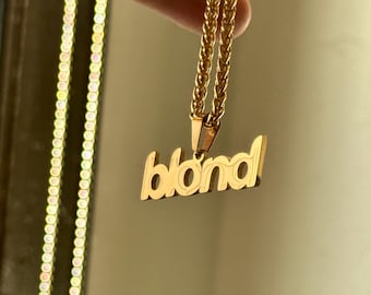 May include: Gold-colored necklace with the word "blond" in a stylized font. The pendant hangs from a delicate chain. The necklace is held up against a neutral background.