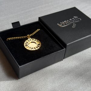 May include: A gold-colored pendant necklace with a sunburst design, displayed in an open black box with a velvet interior. The box has the word "LYRICAL" embossed on the lid.