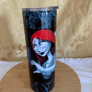 Puede incluir: Un vaso negro y plateado con una imagen de dibujos animados de Sally de The Nightmare Before Christmas. El dise&ntilde;o incluye rosas negras y una Sally pelirroja con rasgos cosidos. El vaso tiene una tapa transparente y est&aacute; dise&ntilde;ado para contener bebidas.