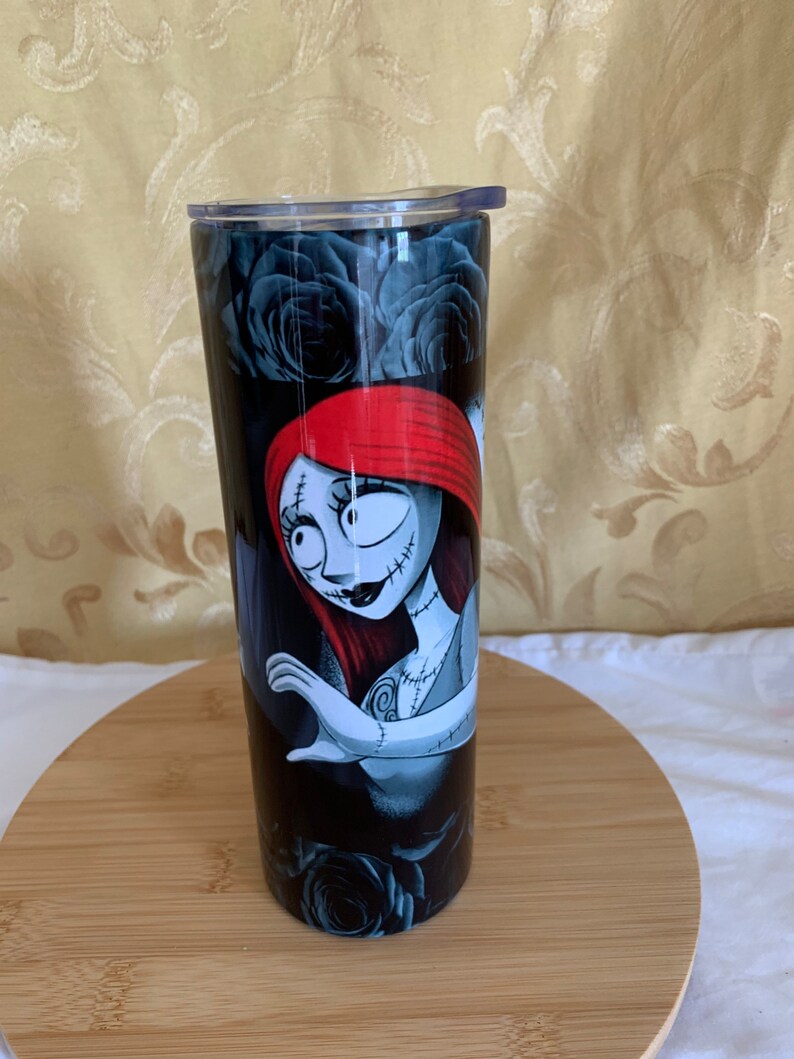 Puede incluir: Un vaso negro y plateado con una imagen de dibujos animados de Sally de The Nightmare Before Christmas. El dise&ntilde;o incluye rosas negras y una Sally pelirroja con rasgos cosidos. El vaso tiene una tapa transparente y est&aacute; dise&ntilde;ado para contener bebidas.