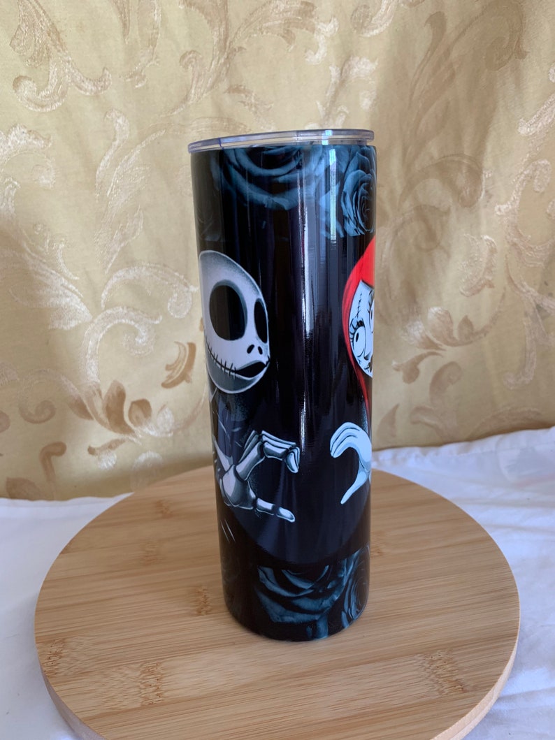 Puede incluir: Un vaso negro con Jack Skellington y Sally de The Nightmare Before Christmas. El dise&ntilde;o incluye rosas negras y un fondo oscuro. El vaso es cil&iacute;ndrico y parece ser de acero inoxidable.