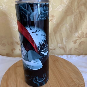 Puede incluir: Un vaso negro con un dise&ntilde;o de Pesadilla antes de Navidad. El dise&ntilde;o incluye a Sally, murci&eacute;lagos y una escena de cementerio. El vaso tiene una tapa plateada y est&aacute; sobre una superficie de madera.