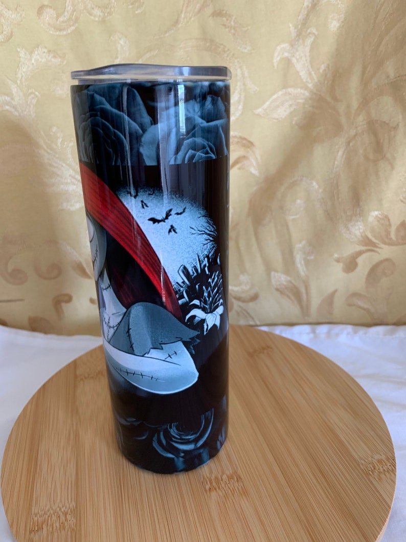 Puede incluir: Un vaso negro con un dise&ntilde;o de Pesadilla antes de Navidad. El dise&ntilde;o incluye a Sally, murci&eacute;lagos y una escena de cementerio. El vaso tiene una tapa plateada y est&aacute; sobre una superficie de madera.