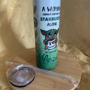 以下が含まれることがあります： 白いタンブラー、蓋とストロー付き。「A WOMAN cannot survive on STARBUCKS ALONE...」の文字と、スターバックスのコーヒーカップを持った緑色のエイリアンのイラストが描かれています。タンブラーは木製のコースターの上にあります。