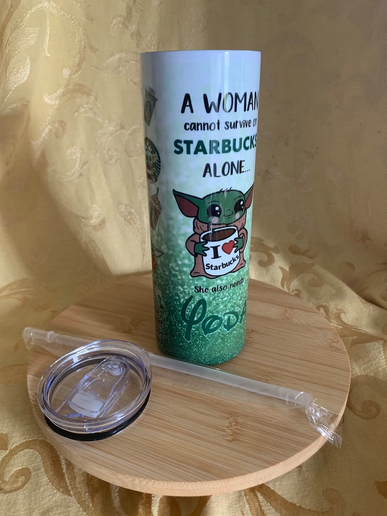 以下が含まれることがあります： 白いタンブラー、蓋とストロー付き。「A WOMAN cannot survive on STARBUCKS ALONE...」の文字と、スターバックスのコーヒーカップを持った緑色のエイリアンのイラストが描かれています。タンブラーは木製のコースターの上にあります。