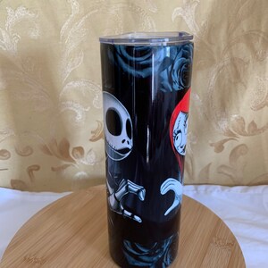Puede incluir: Un vaso negro con Jack Skellington y Sally de Pesadilla antes de Navidad, con un fondo de rosas azul oscuro. El vaso tiene una tapa transparente y se muestra sobre una superficie de madera.