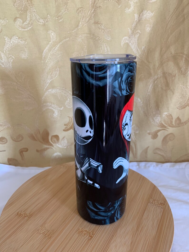 Puede incluir: Un vaso negro con Jack Skellington y Sally de Pesadilla antes de Navidad, con un fondo de rosas azul oscuro. El vaso tiene una tapa transparente y se muestra sobre una superficie de madera.