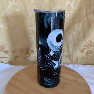 Puede incluir: Un vaso negro y blanco con Jack Skellington de The Nightmare Before Christmas. El dise&ntilde;o incluye rosas negras y una tapa transparente. El vaso es cil&iacute;ndrico y parece estar hecho de acero inoxidable.