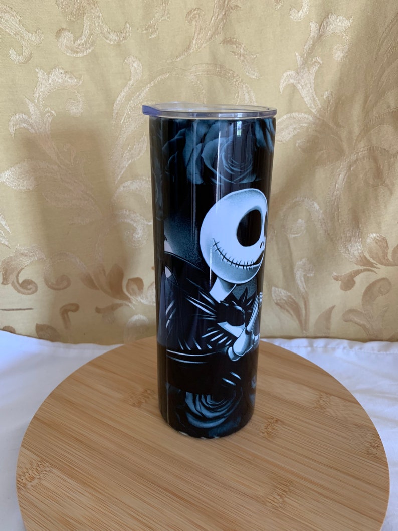 Puede incluir: Un vaso negro y blanco con Jack Skellington de The Nightmare Before Christmas. El dise&ntilde;o incluye rosas negras y una tapa transparente. El vaso es cil&iacute;ndrico y parece estar hecho de acero inoxidable.