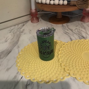 Op de afbeelding: Een groene tumbler met een doorzichtig deksel en de tekst "Speak Fluent Sarcasm" in het wit. De tumbler staat op een gele, geschulpte placemat. De tumbler is cilindrisch en lijkt van metaal te zijn.