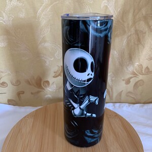 Puede incluir: Un vaso negro con un dise&ntilde;o de Jack Skellington de The Nightmare Before Christmas. El vaso tiene una tapa transparente y est&aacute; decorado con rosas negras. El vaso es cil&iacute;ndrico y se encuentra sobre una superficie de madera.