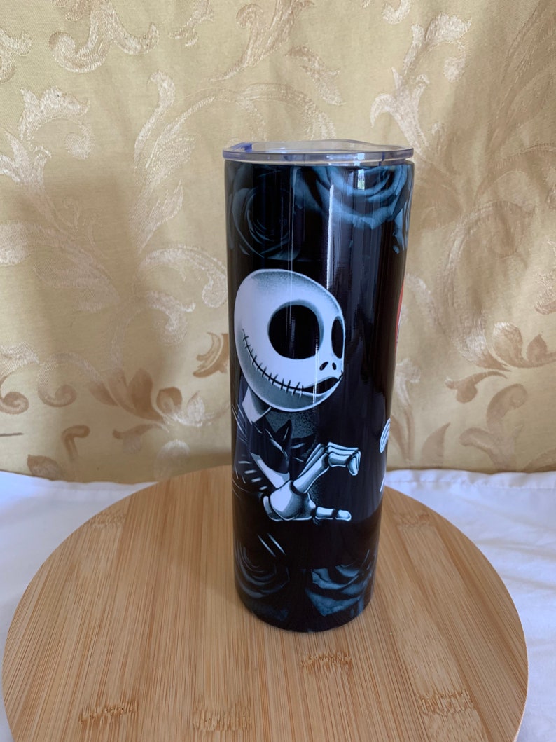 Puede incluir: Un vaso negro con un dise&ntilde;o de Jack Skellington de The Nightmare Before Christmas. El vaso tiene una tapa transparente y est&aacute; decorado con rosas negras. El vaso es cil&iacute;ndrico y se encuentra sobre una superficie de madera.