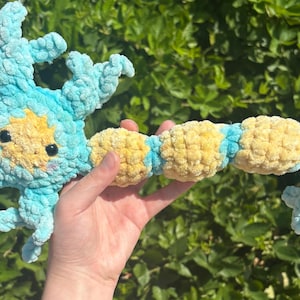 May include: A handmade crochet plush toy in the shape of a sea creature. The toy is primarily light blue with yellow and black accents. The plush has a textured, fuzzy appearance and is held against a background of green foliage.