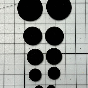 Könnte beinhalten: Eine Anordnung von schwarzen, kreisförmigen Filz-Pads in verschiedenen Größen auf einem Raster. Die Pads haben eine weiche, matte Oberfläche. Geeignet für Bastelarbeiten oder andere kreative Projekte.