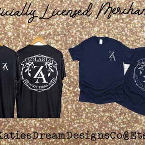 May include: Black and navy blue t-shirts with white graphic designs. The shirts display the letter "A" and the text "SOLARIA AER IGNIS TERRA AQUA". The image also includes the text "Officially Licensed Merchandise" and "KatiesDreamDesignsCo@Etsy".