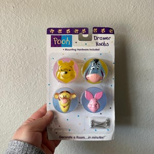 May include: A package of four Winnie the Pooh drawer knobs. Each knob features a different character: Winnie the Pooh, Eeyore, Tigger, and Piglet. The knobs are round with a blue background and include mounting hardware.