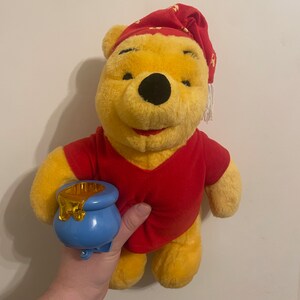 Puede incluir: Un oso de peluche Winnie the Pooh con una camisa de dormir roja y un gorro de dormir rojo y blanco. El oso sostiene un tarro de miel azul con una tapa dorada.