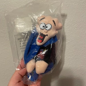 Puede incluir: Un juguete de peluche de un personaje de dibujos animados con una capa azul y la boca muy abierta. El personaje tiene ojos grandes y una expresión de sorpresa.