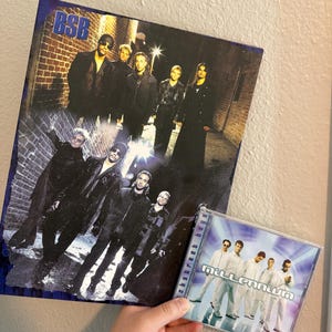 May include: A framed poster and a CD featuring the band Backstreet Boys. The poster has a dark, urban theme with the band members in black clothing. The CD case displays the album title "Millennium" and the band members in white outfits.