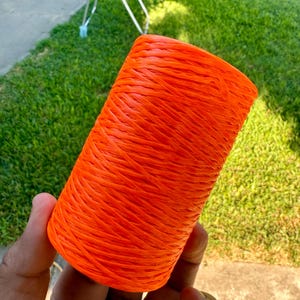 May include: A cylindrical spool of bright orange twine is held in a hand. The twine is tightly wound, showing a textured surface. The background features green grass and a glimpse of a folding chair.