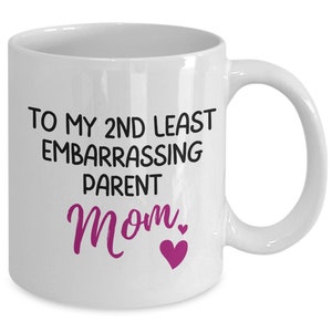 May include: White ceramic mug with the text "TO MY 2ND LEAST EMBARRASSING PARENT Mom" in black and pink lettering. A pink heart graphic is below the word "Mom". The mug has a white handle.