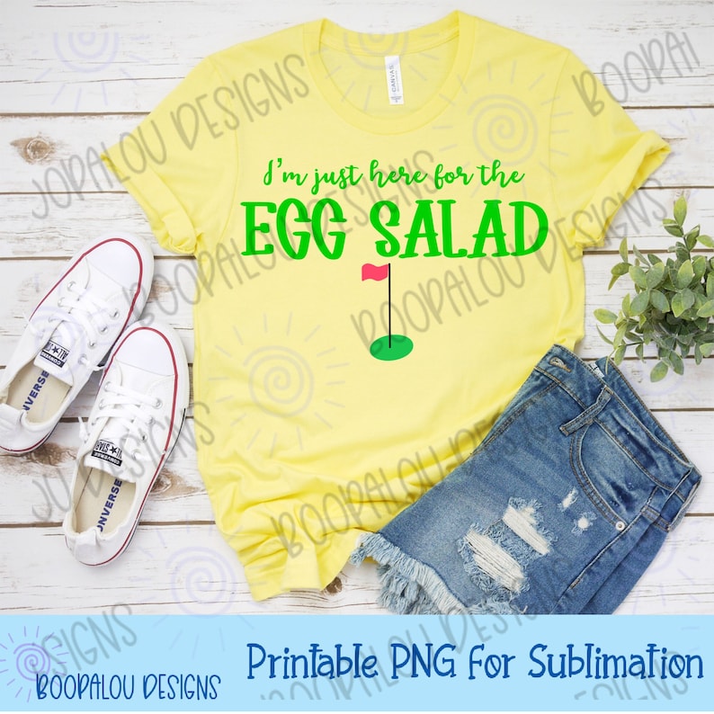 May include: A yellow t-shirt with the text "I'm just here for the EGG SALAD" in green letters. A green golf flag with a red flag is above a green golf hole. The t-shirt is on a wooden surface with a pair of white sneakers and a pair of blue denim shorts.