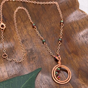 May include: A copper-toned chain necklace featuring a circular pendant made of coiled copper wire. The necklace is accented with small, iridescent, multi-colored beads integrated into the chain and suspended within the center of the circular pendant.