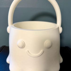 May include: A white, matte-finished bucket shaped like a ghost with a rounded handle. The bucket features a simple, embossed face consisting of two circular eyes and a curved, smiling mouth. The base has a scalloped, wavy edge design.