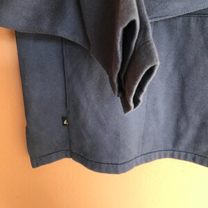 May include: Close-up of a navy blue sweatshirt with a folded sleeve. The sweatshirt has a small, black label with a white logo on the lower left side. The fabric appears to be a soft, knit material.