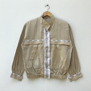 May include: Tan-colored jacket with a stand-up collar and long sleeves. The jacket features a white stripe down the center with text and button closures. The cuffs and waist have elastic bands. The jacket is hanging on a wooden hanger.
