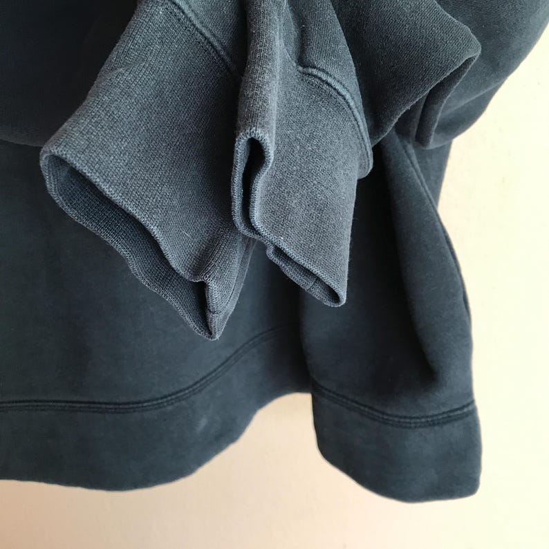 Pu&ograve; includere: Primo piano di una felpa blu scuro. L'immagine mette in evidenza i polsini e l'orlo a coste del capo. Il tessuto appare morbido e leggermente usurato, con dettagli di cucitura visibili. L'estetica generale suggerisce comfort e abbigliamento casual.