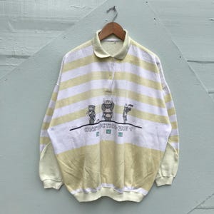 Puede incluir: Camisa de manga larga a rayas amarillas y blancas con cuello y tapeta de botones. La camisa presenta un gráfico de osos de dibujos animados con el texto "CONSTRUCTION ZOE 1".