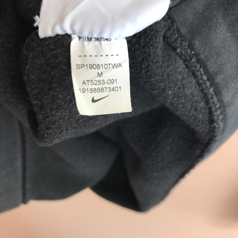 Pu&ograve; includere: Primo piano di un capo di abbigliamento grigio scuro con un'etichetta bianca. L'etichetta presenta il logo Nike e il testo: SP190810TWA, M, AT5253-091 e 19188873401. Il tessuto del capo &egrave; strutturato.