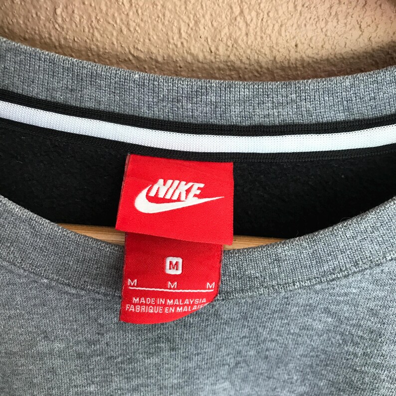 Pu&ograve; includere: Primo piano del colletto di una felpa Nike grigia con interno nero e una striscia bianca. Un'etichetta rossa con il logo Nike e le informazioni sulla taglia &egrave; attaccata. L'etichetta recita "MADE IN MALAYSIA".