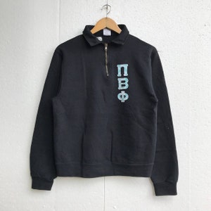 May include: Black sweatshirt with a collared neckline and a quarter-zip closure. The sweatshirt features light blue Greek letters on the front. The long-sleeved top is hanging on a wooden hanger.