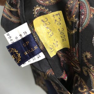 Pu&ograve; includere: Un'etichetta in tessuto blu scuro con il testo "UP HILLS" in lettere dorate. L'etichetta &egrave; attaccata a un tessuto nero con un motivo paisley. Un foglio appiccicoso giallo con il testo "&pound;20.2022 23x23 (24)" &egrave; attaccato al tessuto.