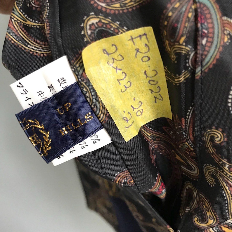 Pu&ograve; includere: Un'etichetta in tessuto blu scuro con il testo "UP HILLS" in lettere dorate. L'etichetta &egrave; attaccata a un tessuto nero con un motivo paisley. Un foglio appiccicoso giallo con il testo "&pound;20.2022 23x23 (24)" &egrave; attaccato al tessuto.