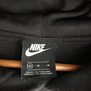 May include: Close-up of a black Nike hoodie label. The label is rectangular with the Nike logo in white. Below the logo are three size indicators, "M", and the text "MADE IN THAILAND FABRIQUE EN THAILANDE".