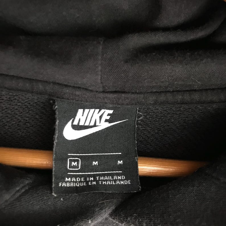 May include: Close-up of a black Nike hoodie label. The label is rectangular with the Nike logo in white. Below the logo are three size indicators, "M", and the text "MADE IN THAILAND FABRIQUE EN THAILANDE".