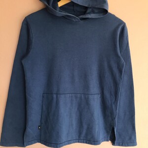 May include: A solid blue hoodie with a kangaroo pocket and a hood. The long-sleeved sweatshirt has a small logo near the bottom hem and a split at the side seams. The garment is made of a soft, casual fabric.
