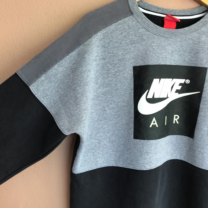 Pu&ograve; includere: Una felpa Nike Air grigia, nera e grigio scuro. La felpa ha un grande quadrato nero con il logo Nike e la scritta "AIR" in bianco. Le maniche sono nere e le spalle grigio scuro.